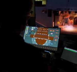 Person operating a lighting console in front of a screen showing stage light controls; lit stage in the background.