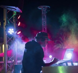 DJ on stage at the pool, working on a laptop; colorful lights and fog create a party vibe.