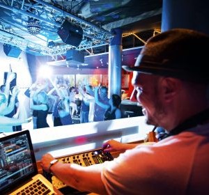 DJ at a mixing console in a club, with people dancing and colorful lights in the background.