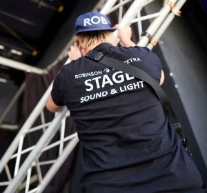 [Translate to Englisch:] Veranstaltungstechniker im Teamoutfit arbeitet an Metallkonstruktion auf der Bühne